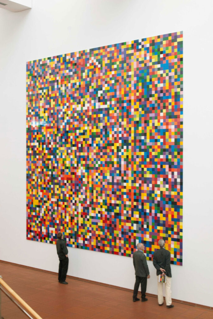 Gerhard Richters Gemälde „4900 Farben“: ein großformatiges Raster aus bunten Farbquadraten an einer weißen Museumswand, davor stehen drei Besucher und betrachten es.